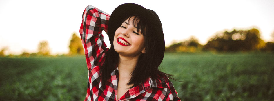 brunette woman wearing black hat and checkered button up, standing outside and smiling