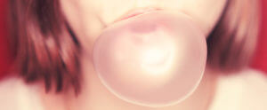 Close up of the mouth of a brunette woman blowing a pink gum bubble against a red wall