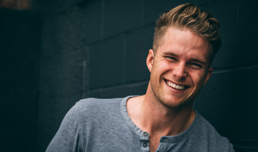 Blonde young man wearing a gray henley smiles while standing against a dark green cinderblock wall