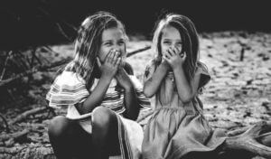 Black and white image of 2 young girls with loose baby teeth laughing and overing their mouths with their hands