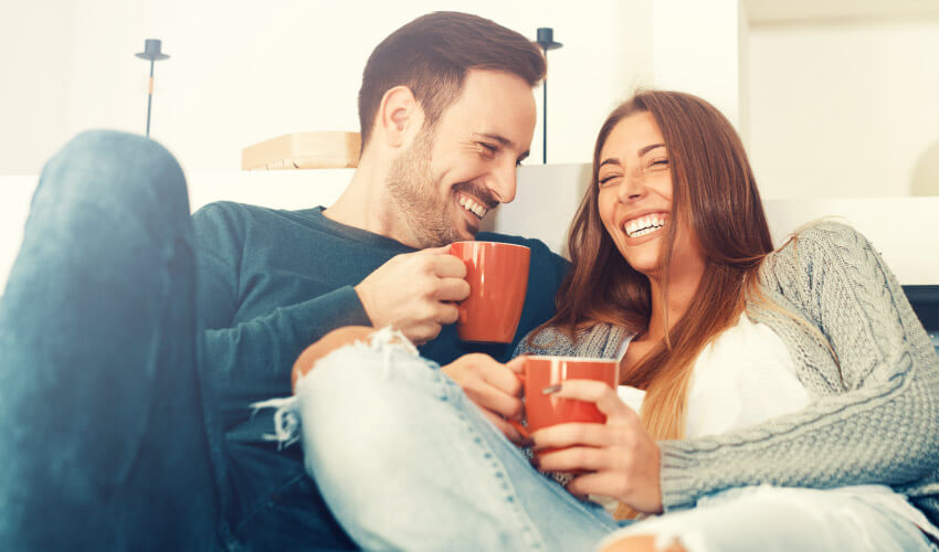 Man and woman smile with dental implants as they cuddle on a couch while holding orange mugs