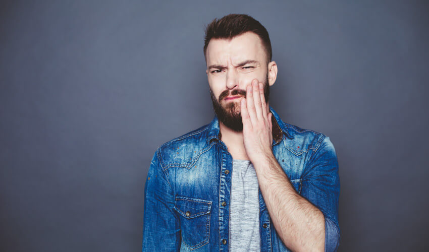 Brunette man with a full beard and mustache cringes and touches his cheek due to pain from a toothache