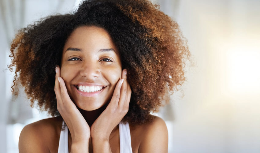 Curly-hair woman touches her cheeks and smiles brightly after easing her dental anxiety