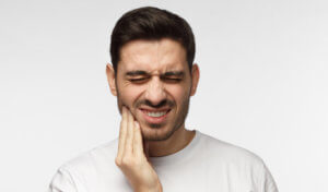 Brunette man in a white t-shirt cringes in pain and touches his cheek due to a toothache dental emergency