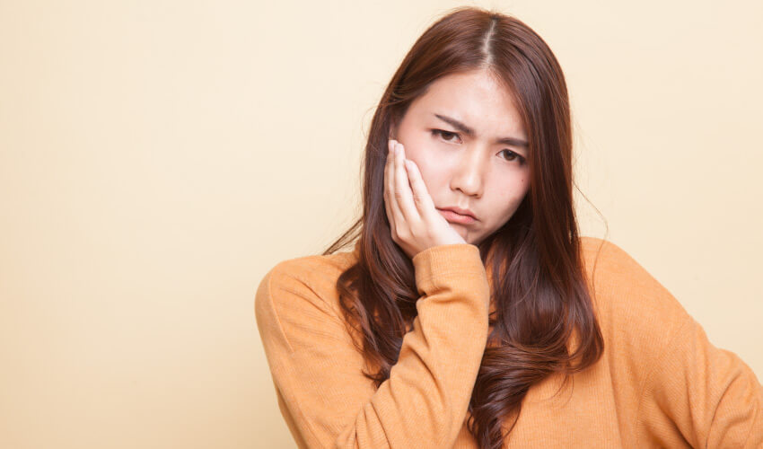 Brunette woman in yellow shirt cringes in pain, cradling her cheek due to a tooth abscess that needs emergency dental care