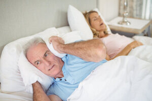 A man covering his ears with his people while his wife snores next to him