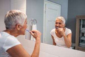 Lady with short gray hair brushes her teeth while looking in the bathroom mirror.