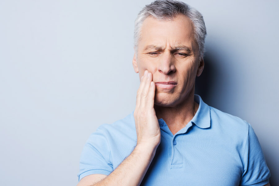 Gray haired man in a blue polo has his hand to his cheek due to pain from a dental abscess.