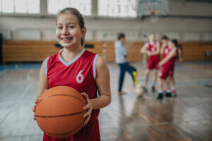 Young tween in braces holding a basketball on the court with teammates in the background.